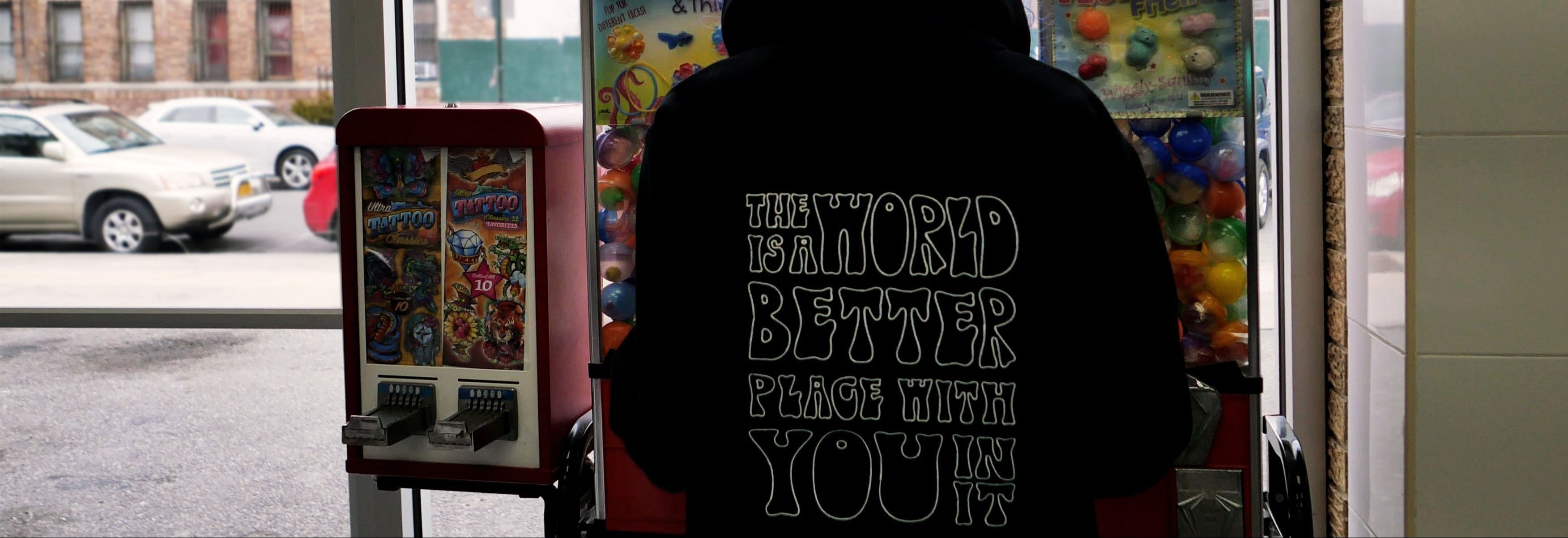 Person wearing a hoodie with text in front of a vending machine on a city street.