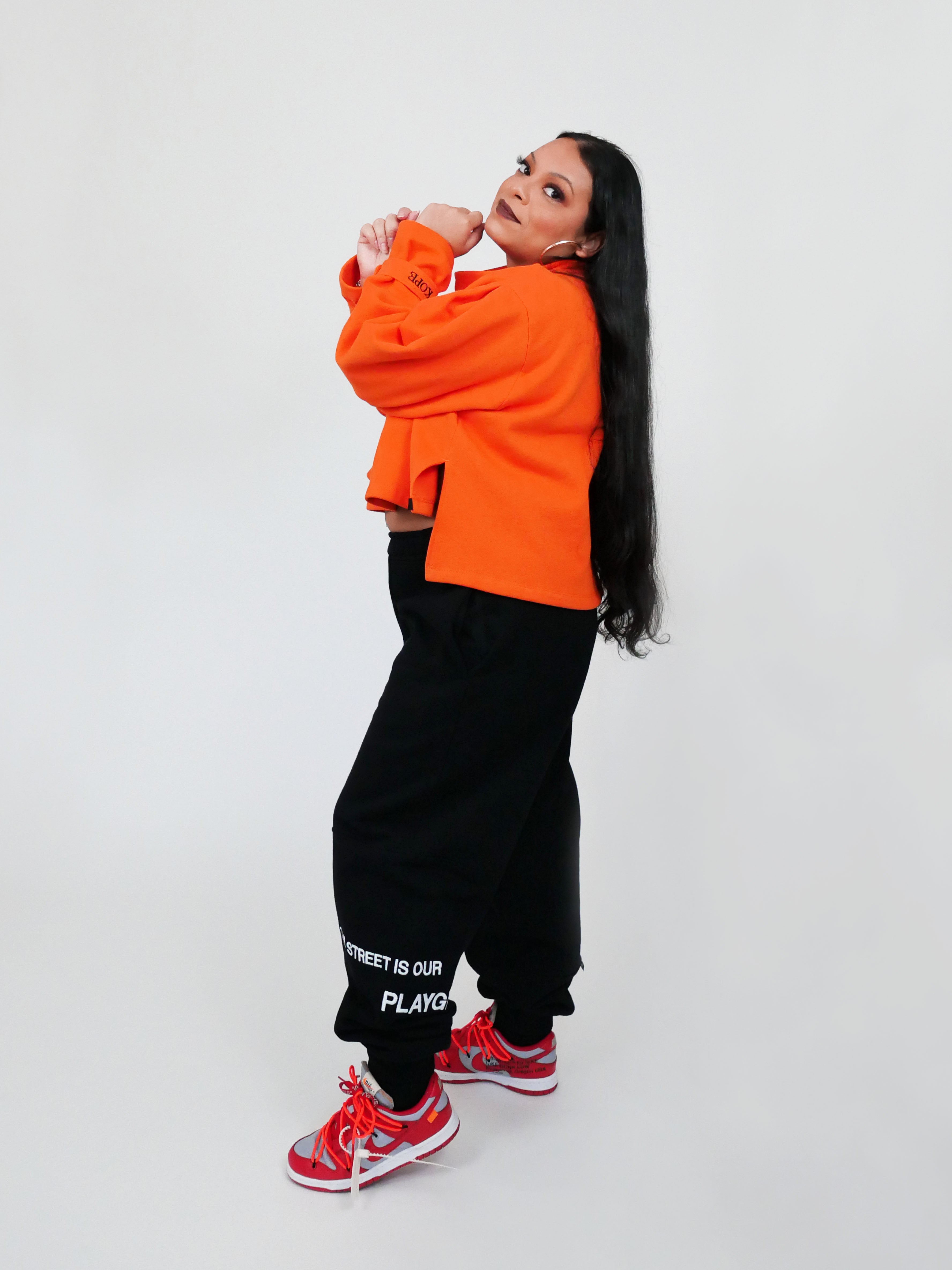 Person wearing an orange sweatshirt and black sweatpants with text, standing against a white background