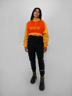 Person wearing a two-tone orange and yellow half zip-up with a logo design, black sweatpants, and black boots on a light gray background