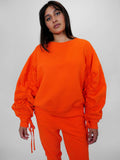 Person wearing a bright orange crewneck with ruched sleeves on a plain background