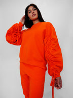 Person wearing an orange crewneck with ruched sleeves on a plain background
