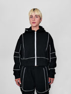 Person wearing a black full zip hoodie and sweatpants with white piping on a plain background