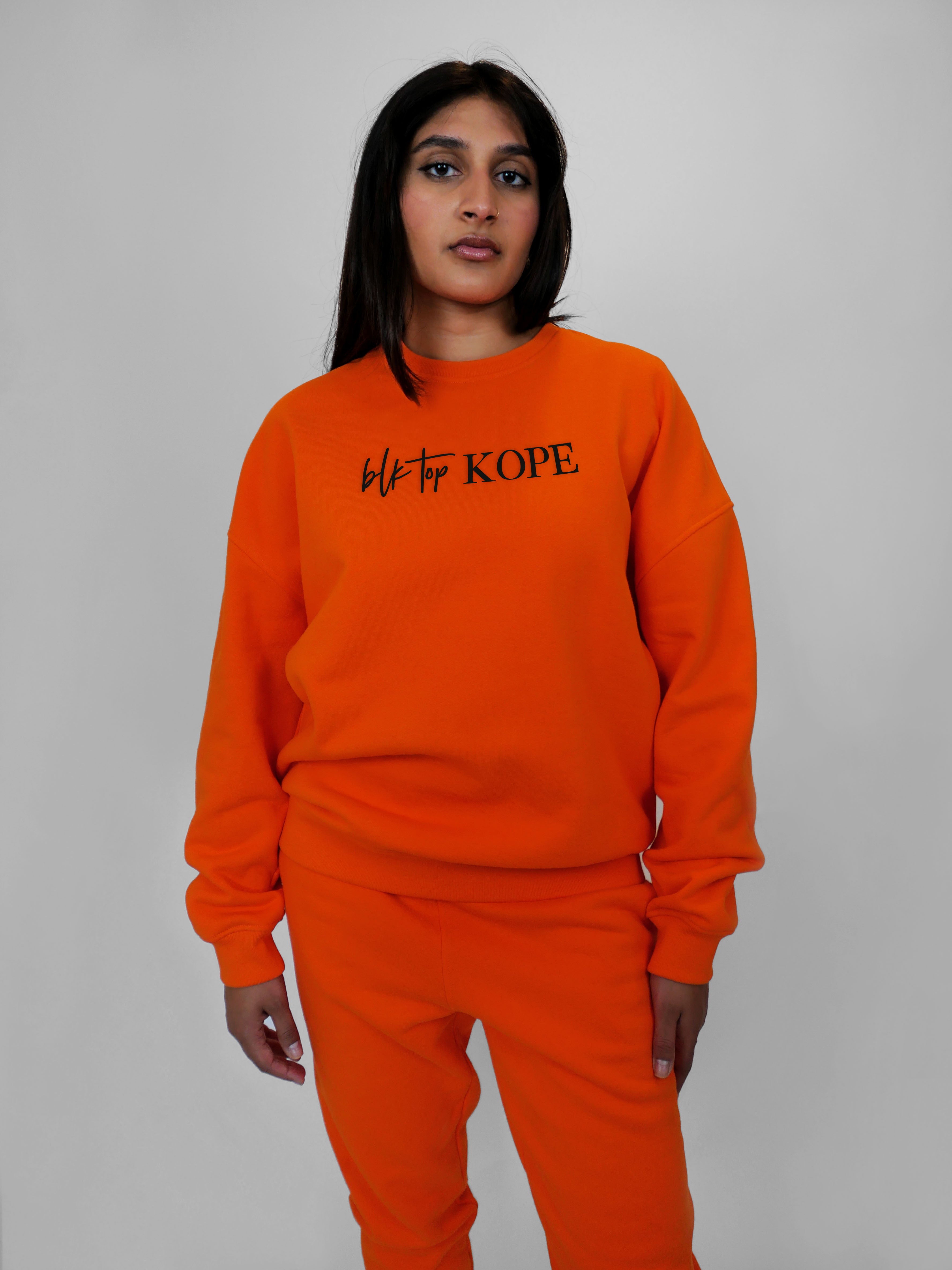 Person wearing an orange crewneck with 'blk top KOPE' text on a gray background