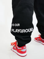 Black sweatpants with 'THE STREET IS OUR PLAYGROUND' text worn by a person with red sneakers on a white background