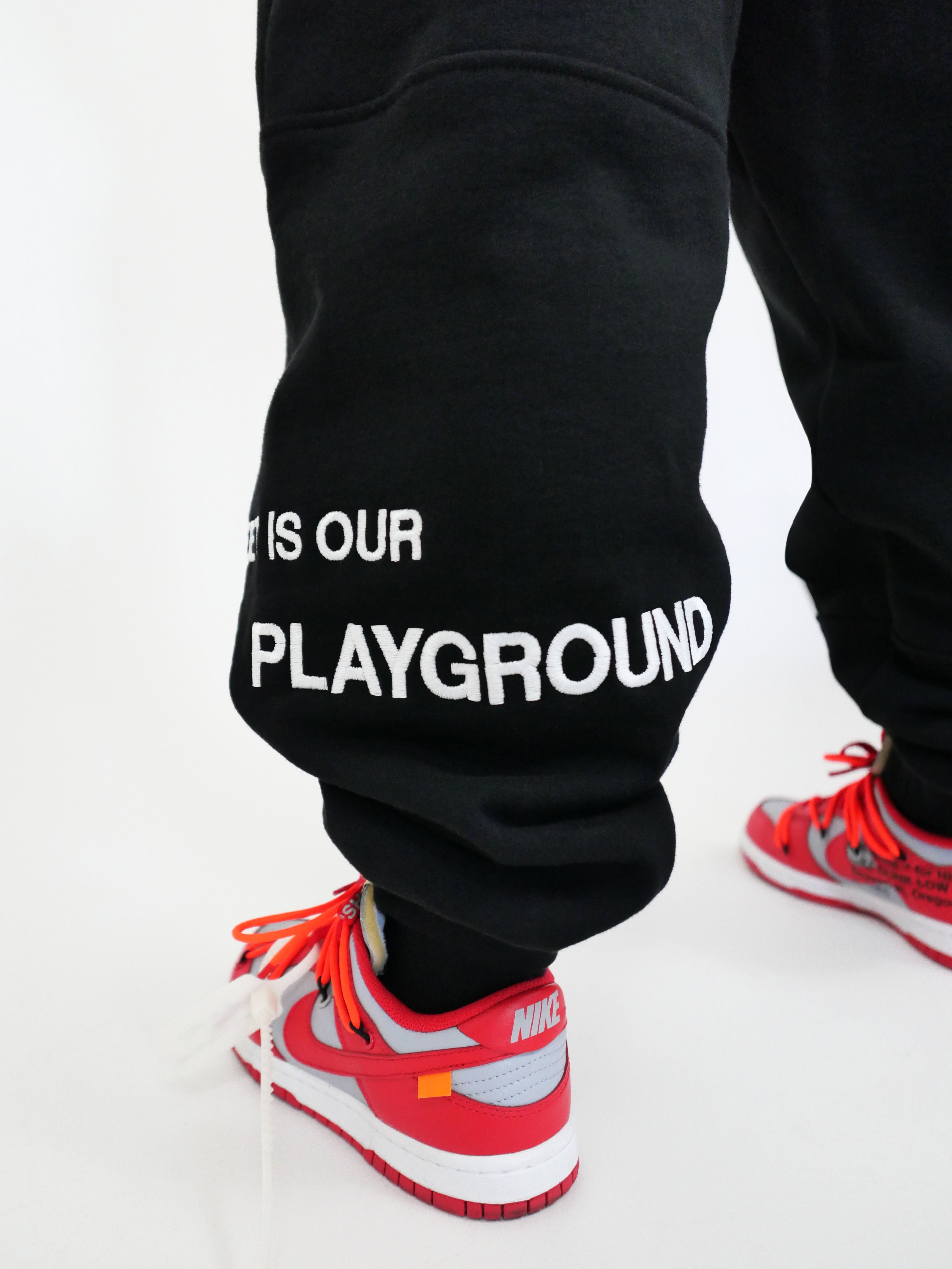 Black sweatpants with 'THE STREET IS OUR PLAYGROUND' text worn by a person with red sneakers on a white background