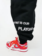 Black sweatpants with 'The street is our playground' text and red sneakers on a white background