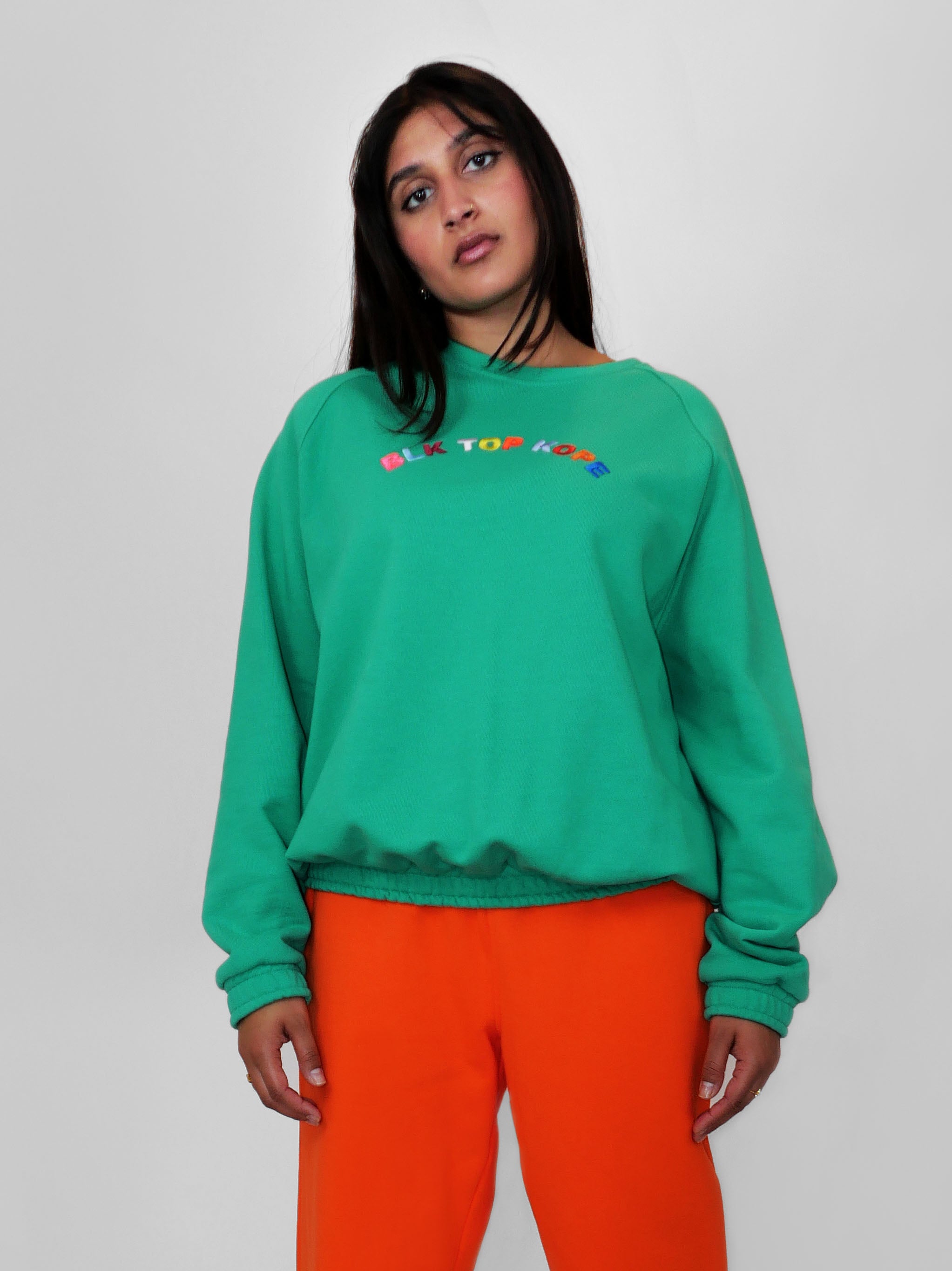 Woman wearing a green crewneck with colorful text and orange pants on a light gray background