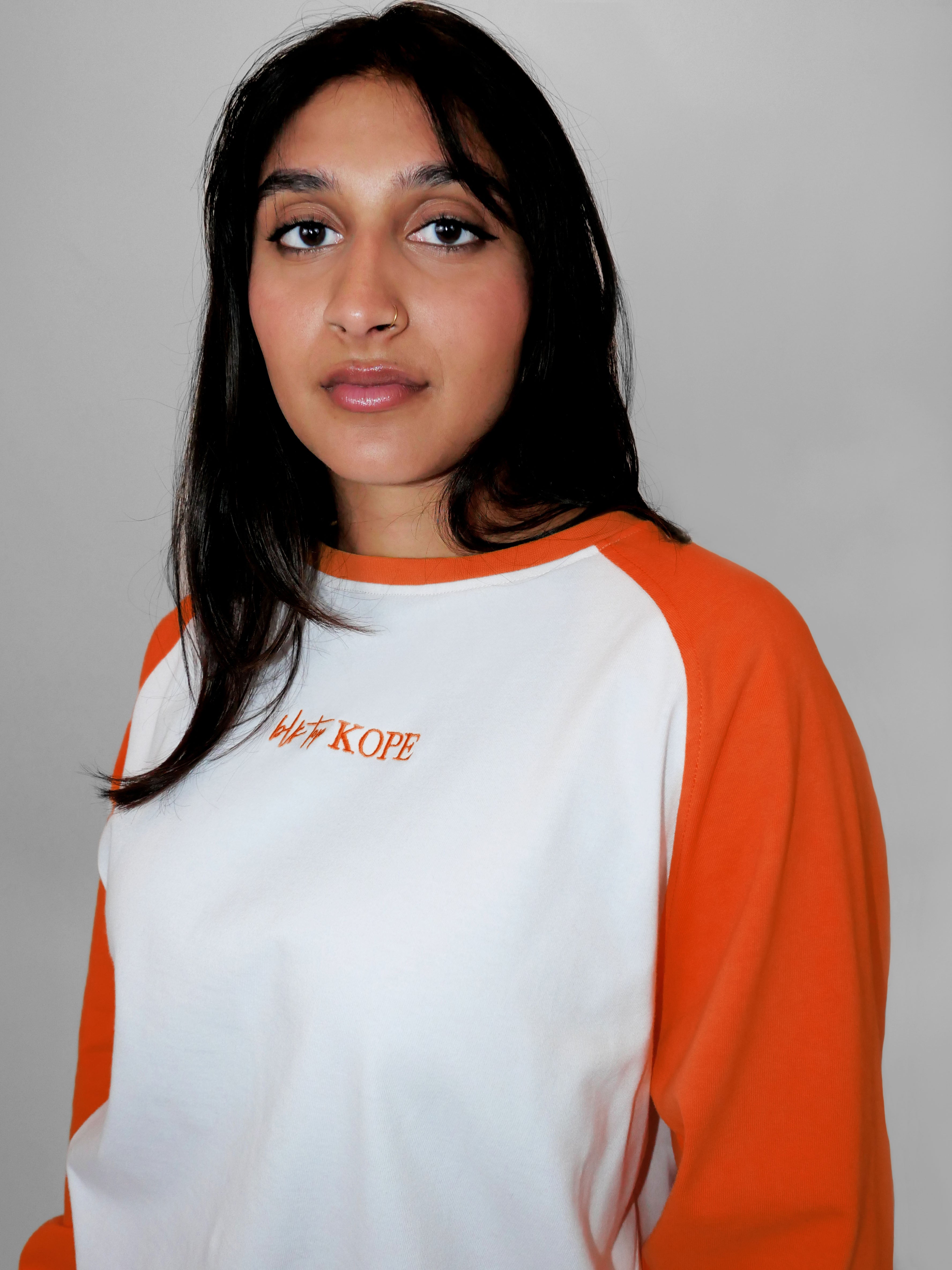 Woman wearing an orange and white raglan longsleeve with a brand logo on a plain background