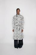 A model stands against a light gray studio background wearing a long-sleeved, high-neck trenchcoat with an all-over black and white abstract pattern, paired with loose black pants.