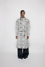 A model stands against a light gray studio background wearing a long-sleeved, high-neck trenchcoat with an all-over black and white abstract pattern, paired with loose black pants.