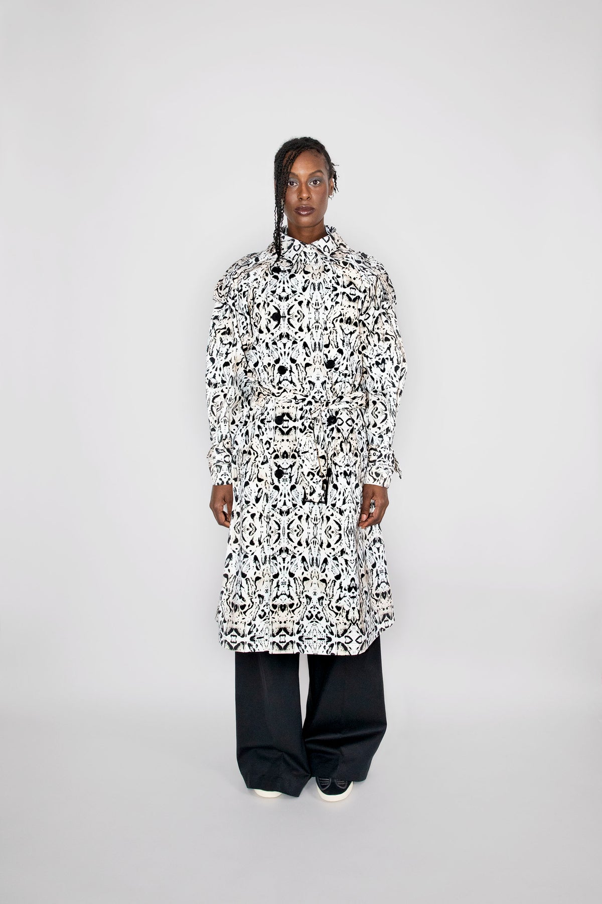 A model stands against a light gray studio background wearing a long-sleeved, high-neck trenchcoat with an all-over black and white abstract pattern, paired with loose black pants.