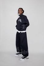 A model wearing a black track jacket with white graphic embroidery 'New York', matching black wide-leg pants with flame embroidery near the hem, and black low-top sneakers with white soles, posed against a light gray studio background.