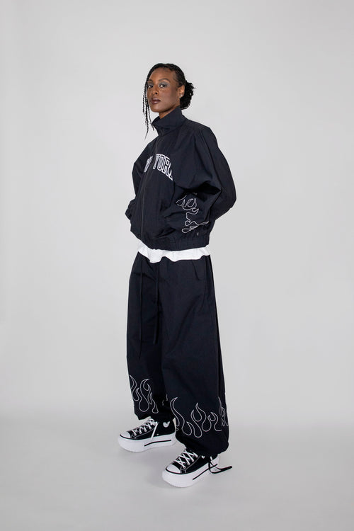 A model wearing a black track jacket with white graphic embroidery 'New York', matching black wide-leg pants with flame embroidery near the hem, and black low-top sneakers with white soles, posed against a light gray studio background.