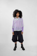 A model with curly black hair stands against a light gray studio background wearing a light purple mesh long-sleeve top over a lavender shirt, dark cropped shorts, and black boots.