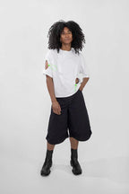A model with curly black hair stands against a light gray studio background wearing a white short-sleeve t-shirt with lime green graphic details, black wide-leg cropped shorts, and black ankle boots.