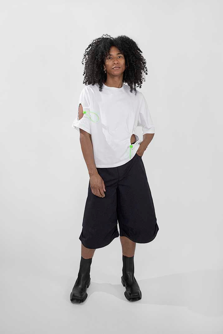 A model with curly black hair stands against a light gray studio background wearing a white short-sleeve t-shirt with lime green graphic details, black wide-leg cropped shorts, and black ankle boots.