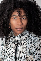 A close-up portrait of a person with voluminous curly black hair wearing a high-neck zip-up jacket with a black and white abstract organic pattern.