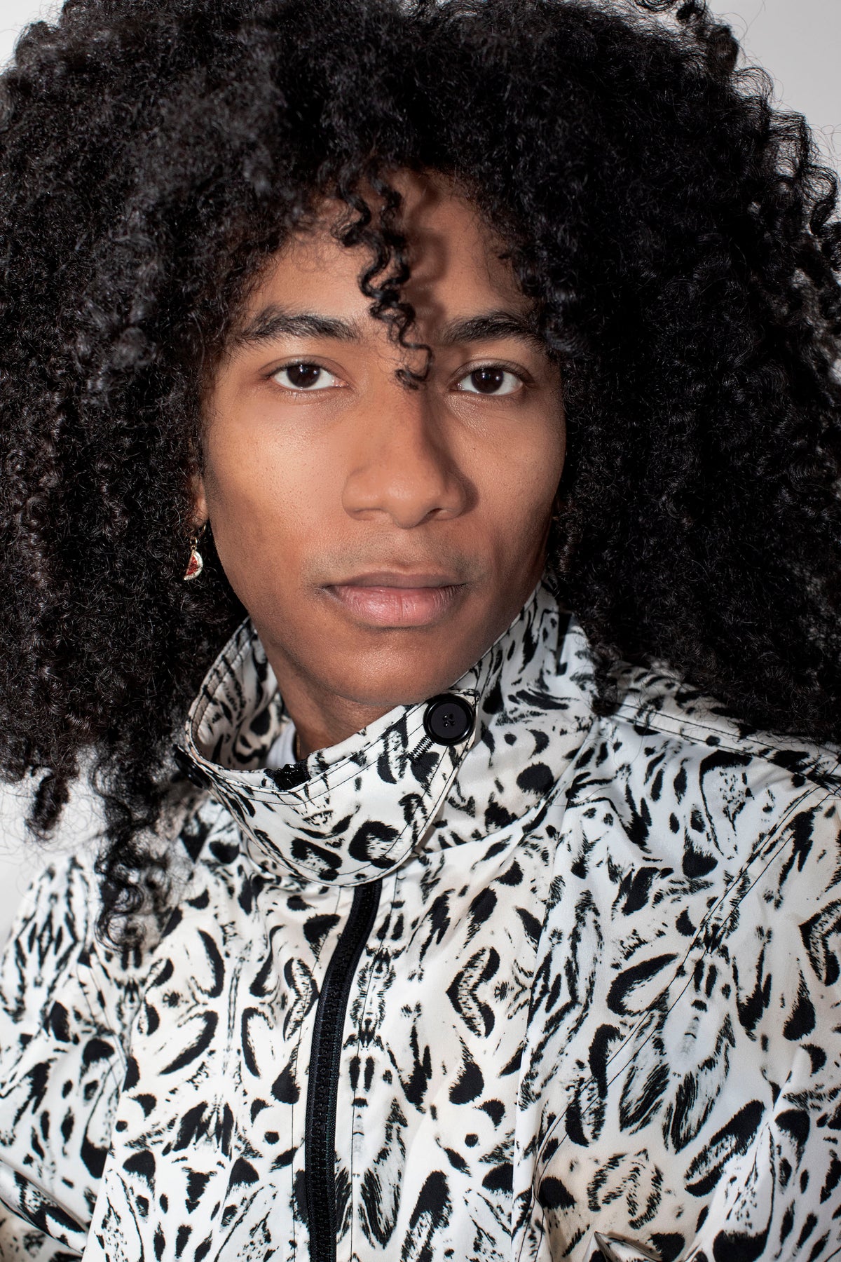 A close-up portrait of a person with voluminous curly black hair wearing a high-neck zip-up jacket with a black and white abstract organic pattern.