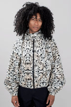 A model with voluminous black curly hair wearing a black and off-white abstract patterned zip-up jacket and dark trousers, photographed against a neutral light gray background.