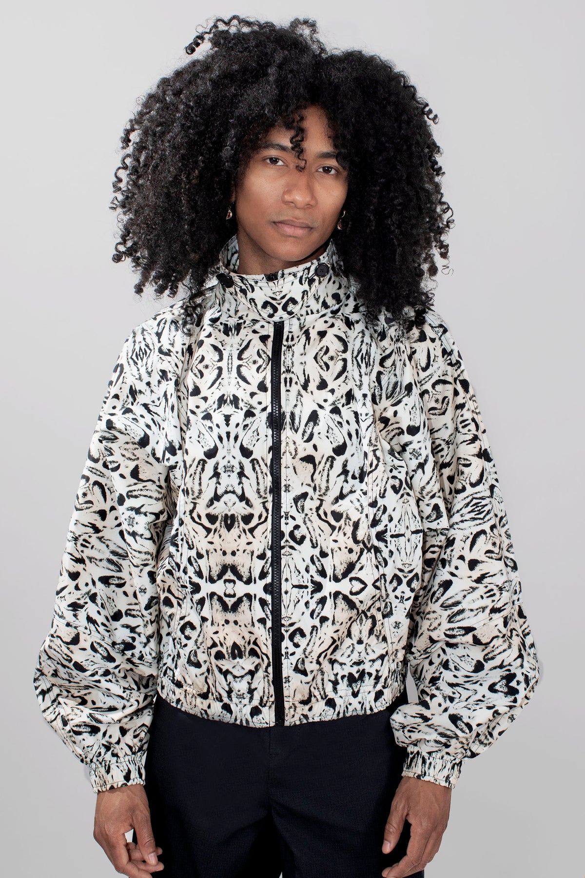 A model with voluminous black curly hair wearing a black and off-white abstract patterned zip-up jacket and dark trousers, photographed against a neutral light gray background.