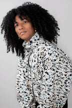 A person with voluminous black curly hair wearing a high-neck zip-up track jacket with a black and white abstract organic pattern, photographed against a plain light gray background.