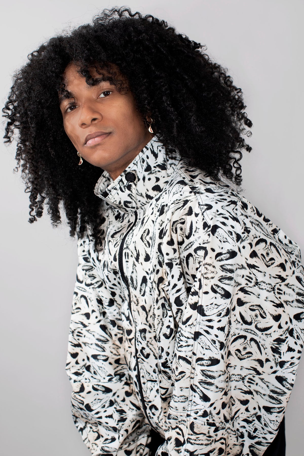 A person with voluminous black curly hair wearing a high-neck zip-up track jacket with a black and white abstract organic pattern, photographed against a plain light gray background.