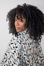 A person with voluminous black curly hair wearing a balloon track jacket with a black and white abstract animal print, photographed against a plain light gray background