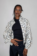 A model wearing a black and white abstract patterned balloon track jacket over a black crop top and dark trousers, photographed against a neutral light gray studio background.