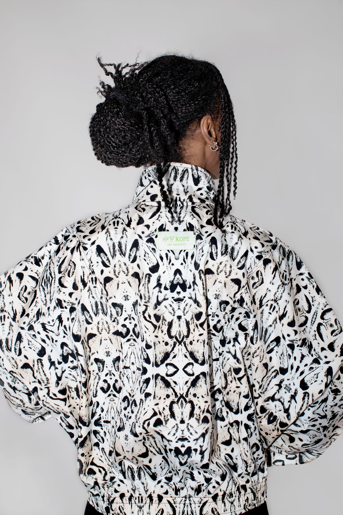 A person shown from the back wearing a high-neck, voluminous jacket with an all-over black and white abstract symmetrical pattern, set against a plain light gray background.