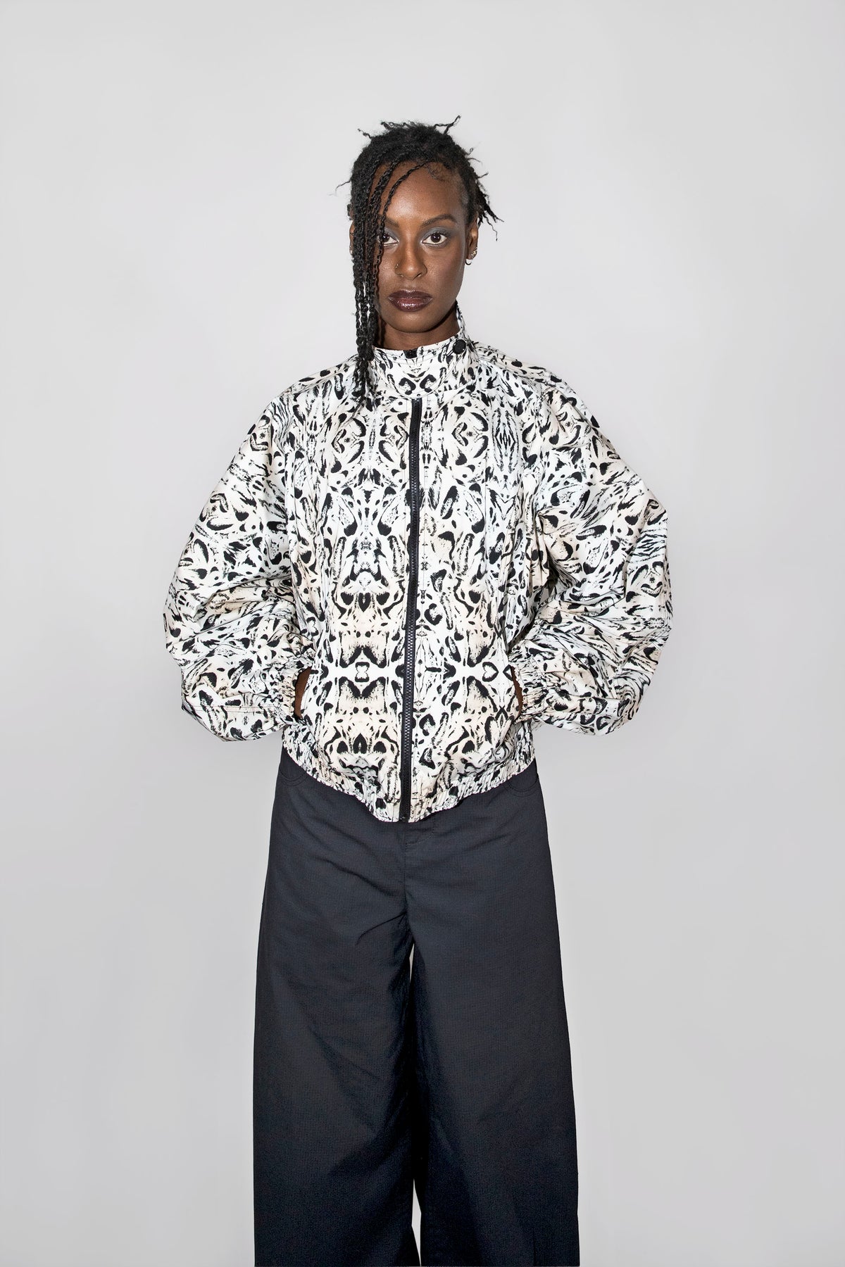 A model stands against a light gray studio background wearing a balloon track jacket with a black-and-white abstract pattern and dark wide-leg pants.