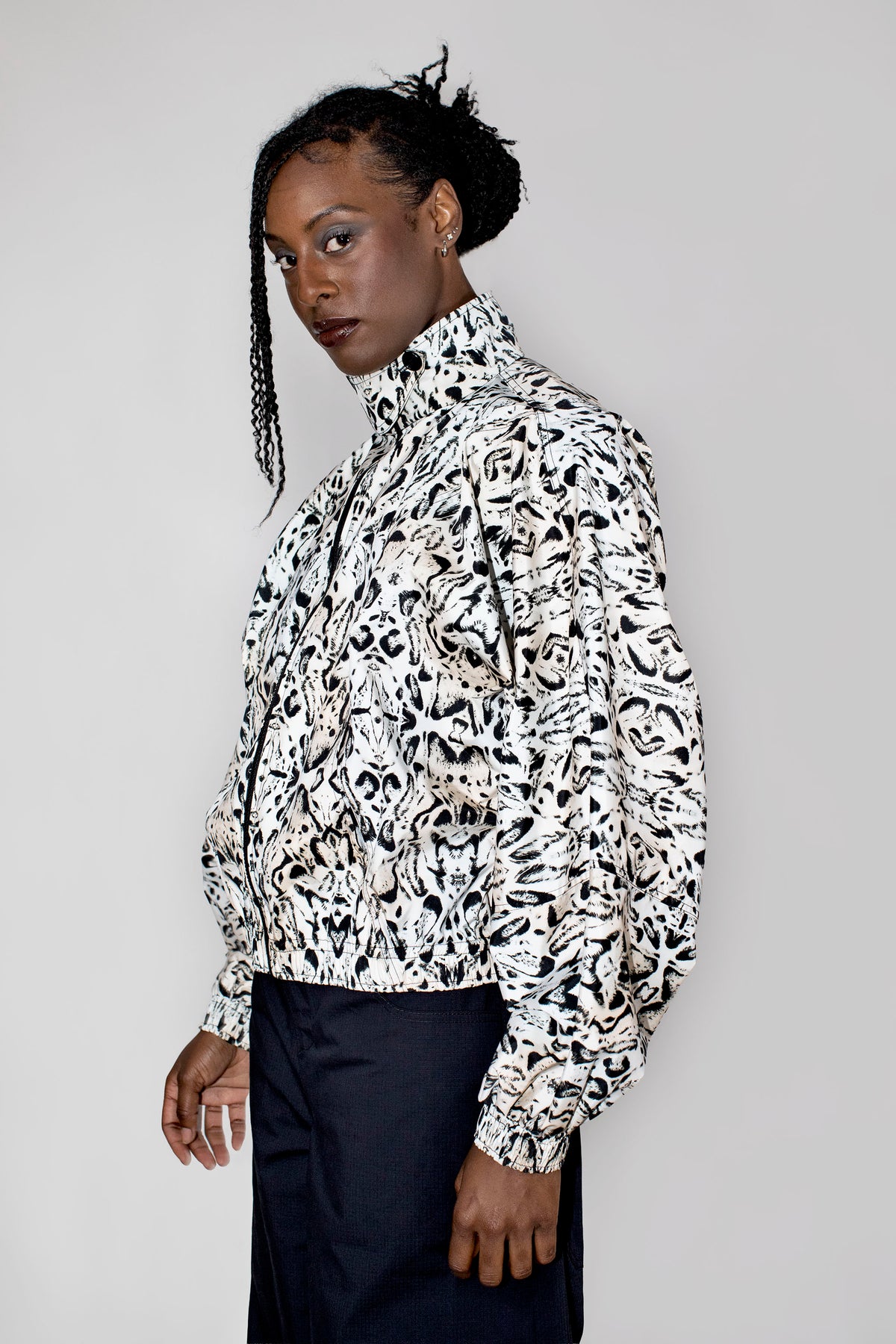 A model wearing a black and white abstract patterned high-neck track jacket with dark trousers, photographed against a solid light gray studio background.