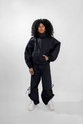 A model with curly black hair stands against a light gray studio background wearing a dark oversized matching track jacket and pants set with white sneakers.