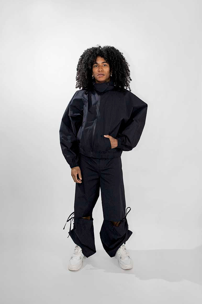 A model with curly black hair stands against a light gray studio background wearing a dark oversized matching track jacket and pants set with white sneakers.