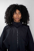 A person with voluminous curly black hair wearing a black high-collar zip-front track jacket, photographed against a plain light gray background.