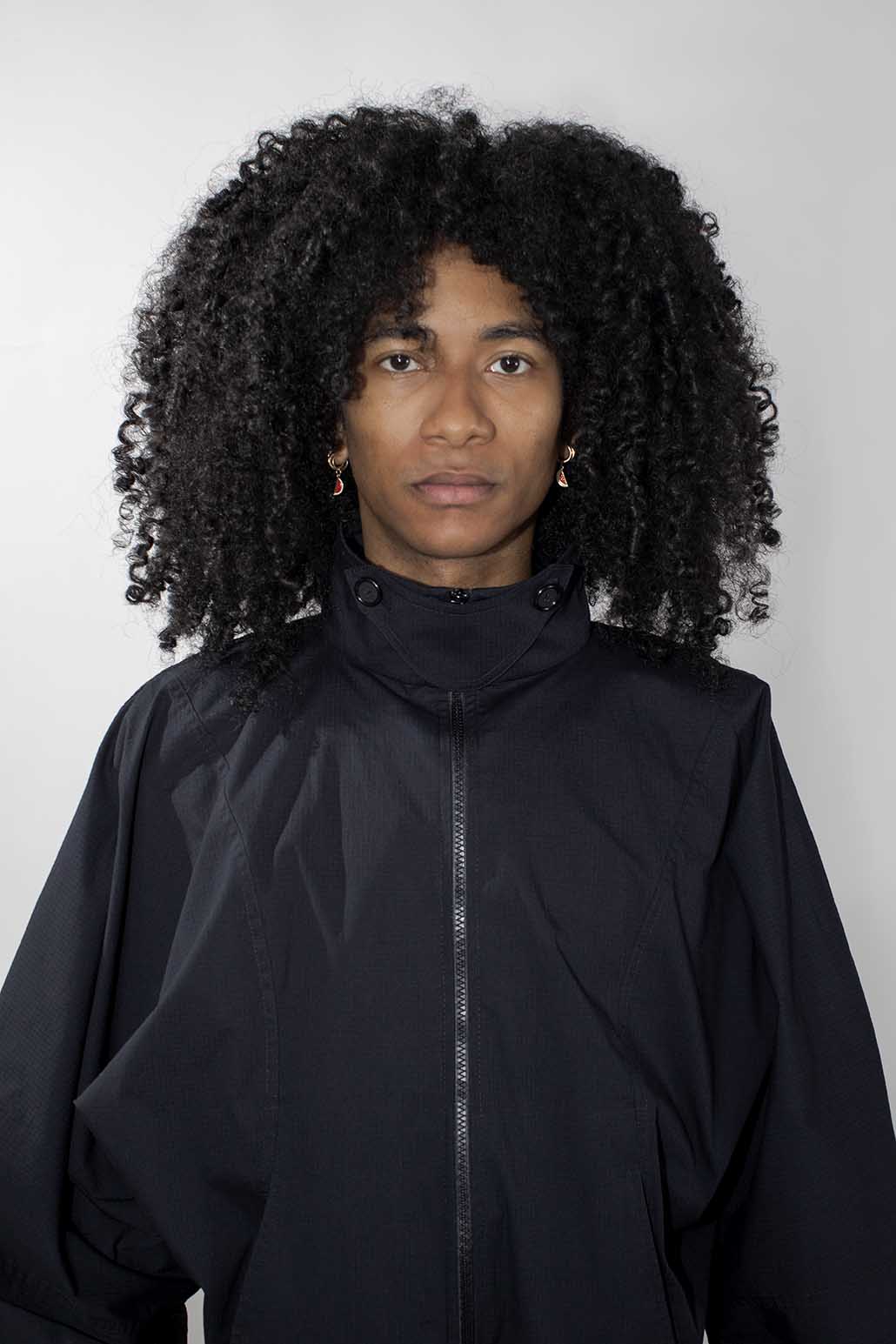 A person with voluminous curly black hair wearing a black high-collar zip-front track jacket, photographed against a plain light gray background.