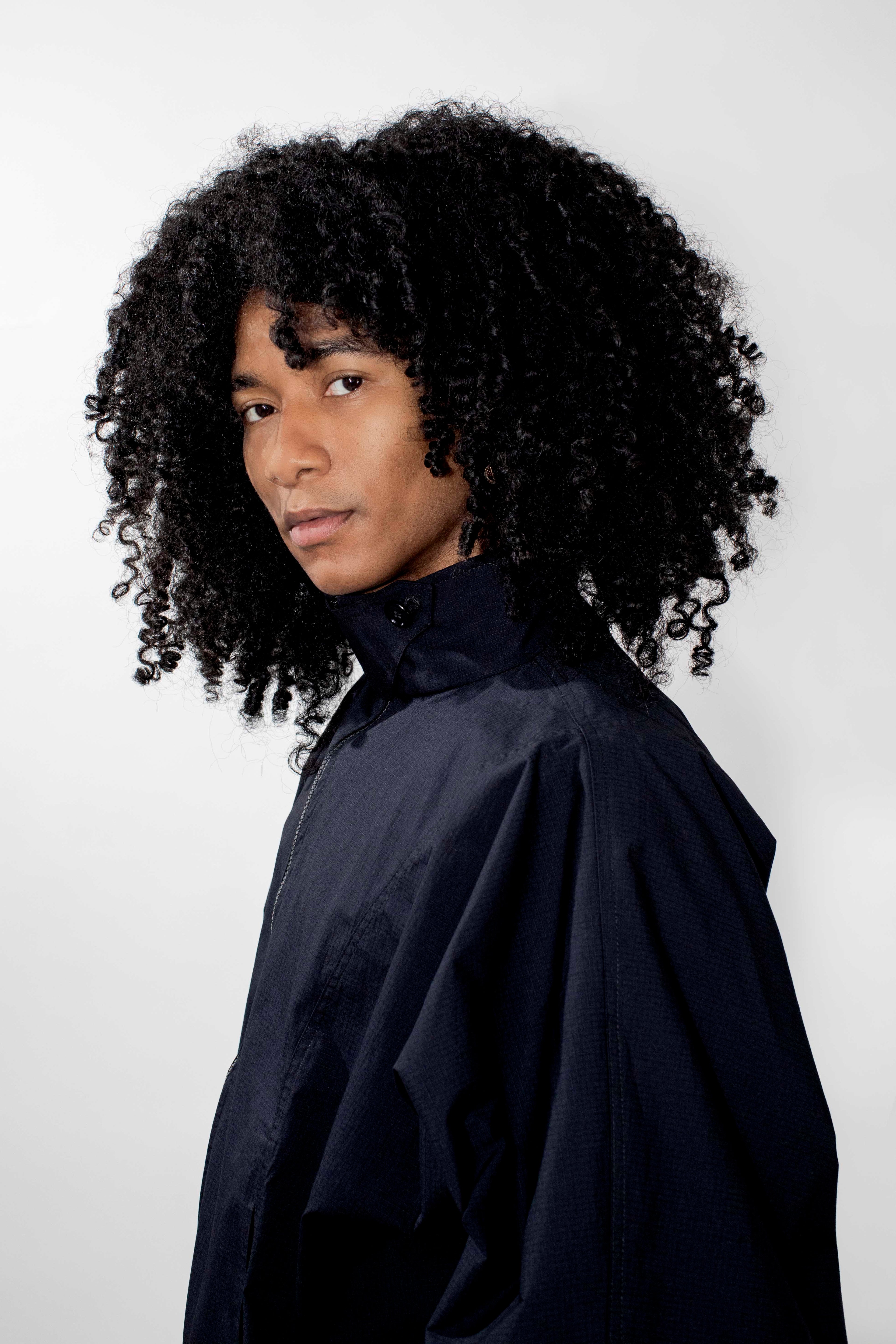 A person with voluminous dark curly hair wearing a dark high-neck track jacket, photographed against a plain light gray studio background.
