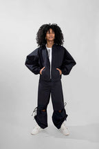 A model with curly black hair stands against a light gray studio background wearing a dark oversized track zip-front jacket, matching loose pants, a white top, and white sneakers.