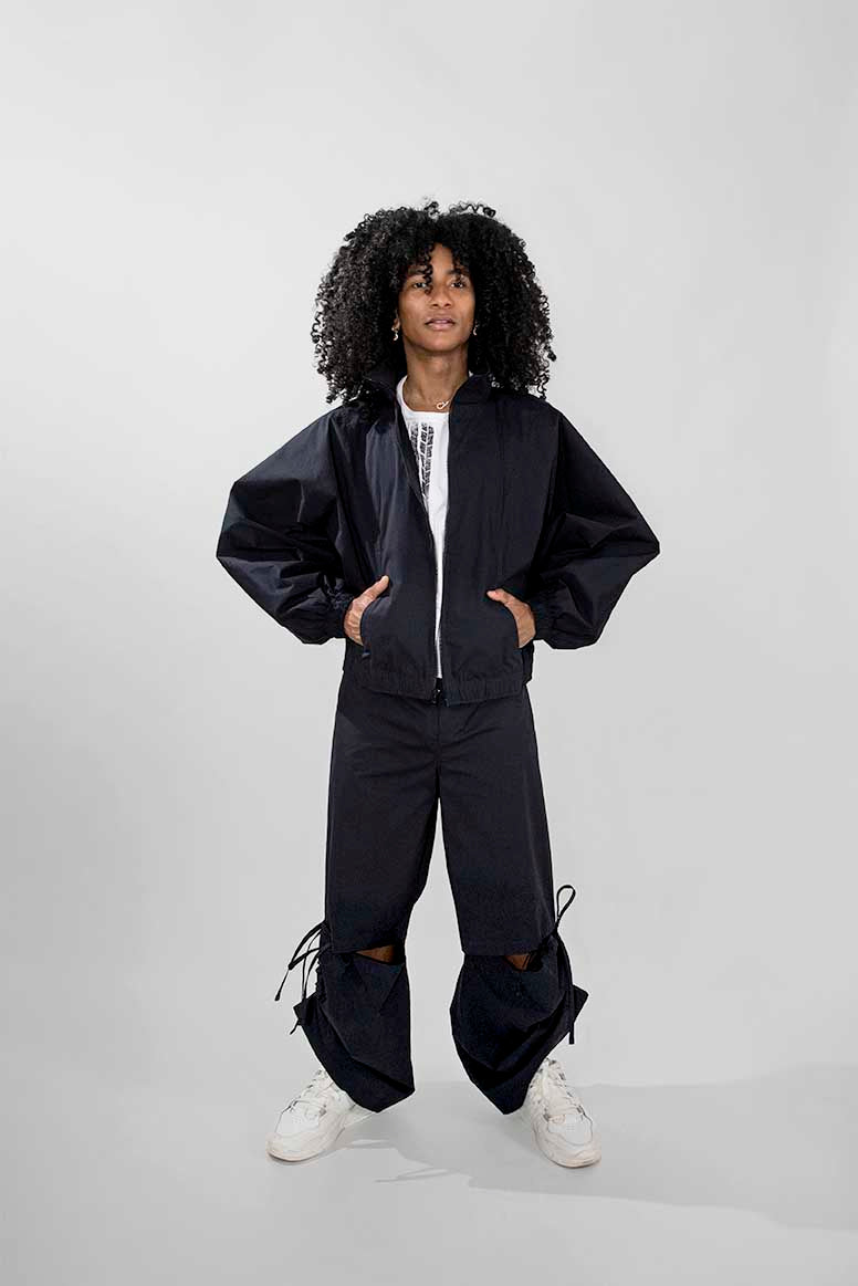 A model with curly black hair stands against a light gray studio background wearing a dark oversized track zip-front jacket, matching loose pants, a white top, and white sneakers.