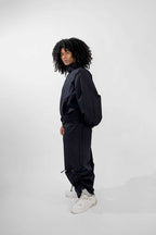 A model with curly black hair stands in profile wearing a black oversized track jacket and matching loose pants, paired with white sneakers, against a light gray studio background.