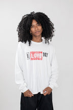 A model with curly black hair wearing a white long-sleeve t-shirt with a red and black 'I LOVE NY' graphic on the chest, paired with black pants, against a light gray background.