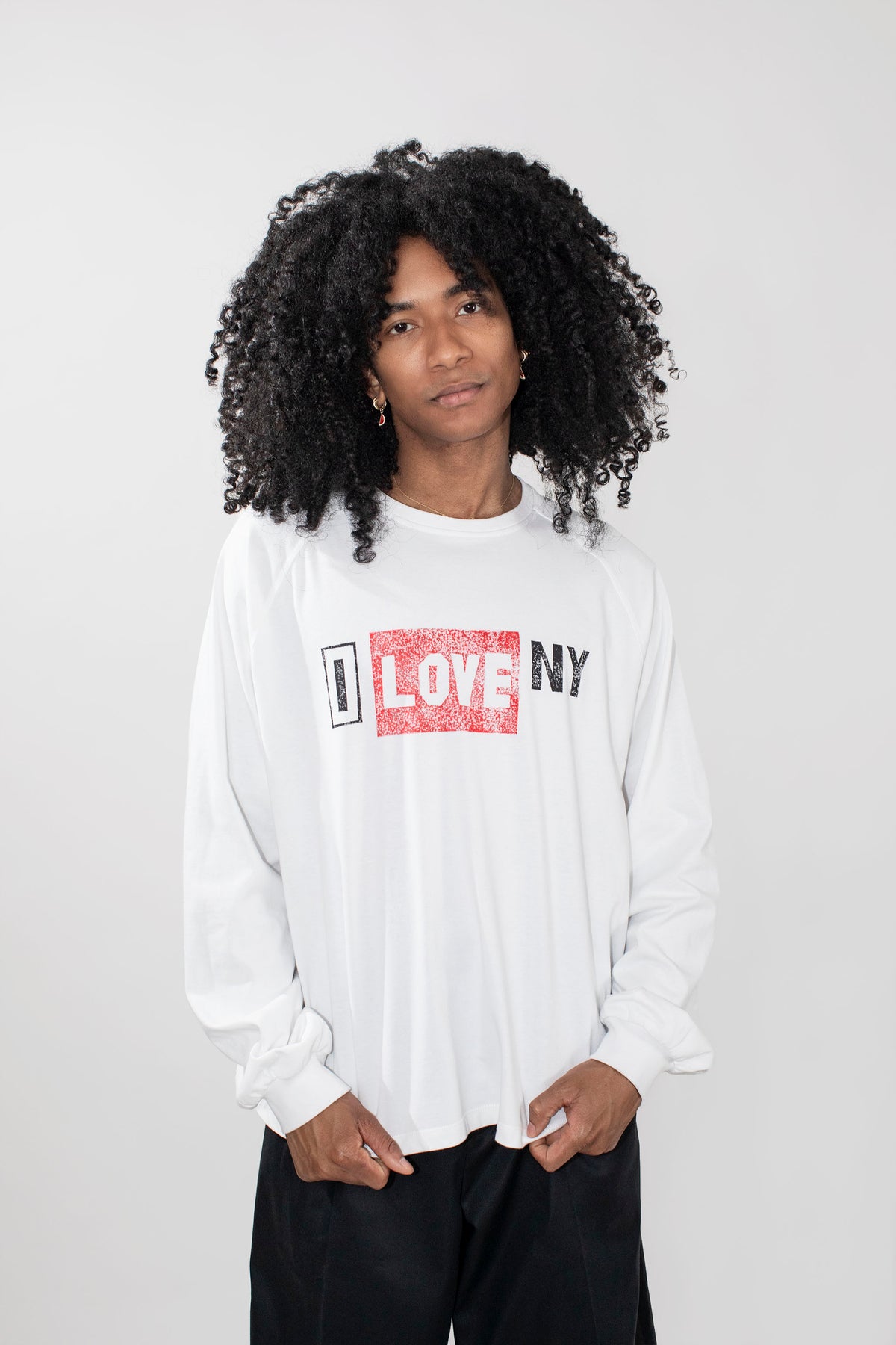 A model with curly black hair wearing a white long-sleeve t-shirt with a red and black 'I LOVE NY' graphic on the chest, paired with black pants, against a light gray background.