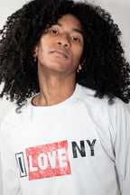 A close-up shot of a person with voluminous black curly hair wearing a white long-sleeve t-shirt with a graphic print that reads 'I LOVE NY' in red and black.