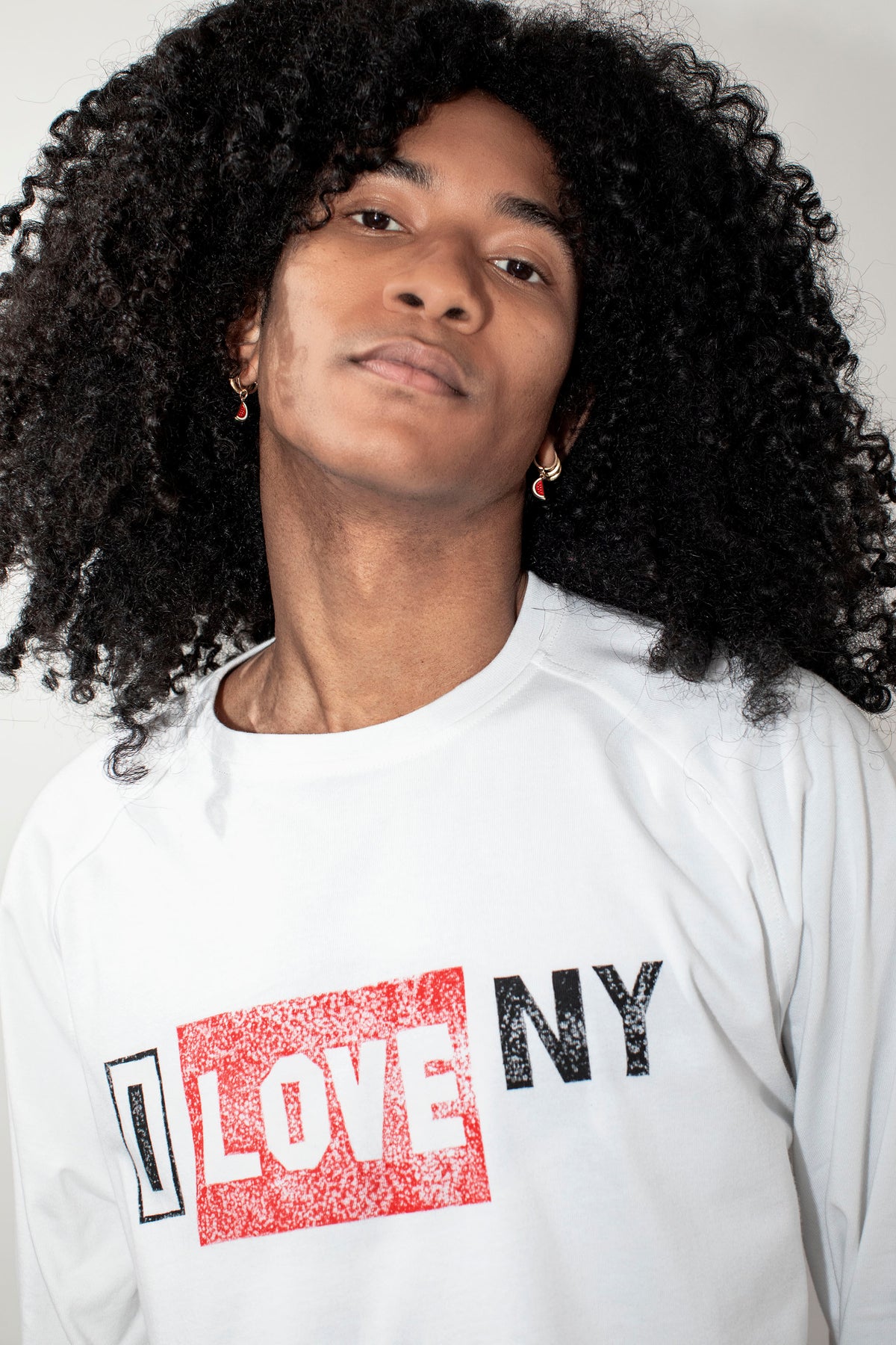A close-up shot of a person with voluminous black curly hair wearing a white long-sleeve t-shirt with a graphic print that reads 'I LOVE NY' in red and black.
