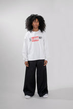 A model with curly black hair stands against a light gray studio background wearing a white long-sleeve crewneck sweatshirt with a red and black 'I LOVE NY' graphic on the chest, paired with black wide-leg sweatpants and white sneakers.