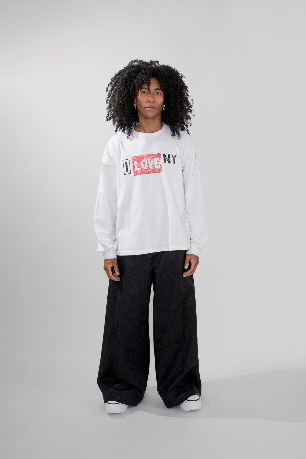 A model with curly black hair stands against a light gray studio background wearing a white long-sleeve crewneck sweatshirt with a red and black 'I LOVE NY' graphic on the chest, paired with black wide-leg sweatpants and white sneakers.