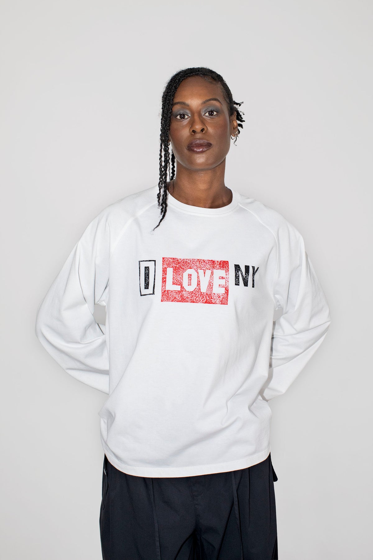 A model wearing a white long-sleeve sweatshirt with a 'I LOVE NY' graphic on the chest, paired with black pants, against a plain light gray background.