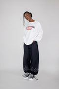 A model stands against a light gray studio background wearing a white long-sleeve crewneck sweatshirt with a red and black 'I LOVE NY' graphic, black wide-leg sweatpants with white flame graphics near the ankles, and black-and-white sneakers.