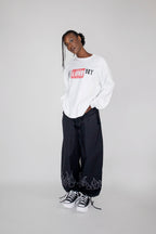 A model stands against a light gray studio background wearing a white long-sleeve crewneck sweatshirt with a red and black 'I LOVE NY' graphic, black wide-leg sweatpants with white flame graphics near the ankles, and black-and-white sneakers.