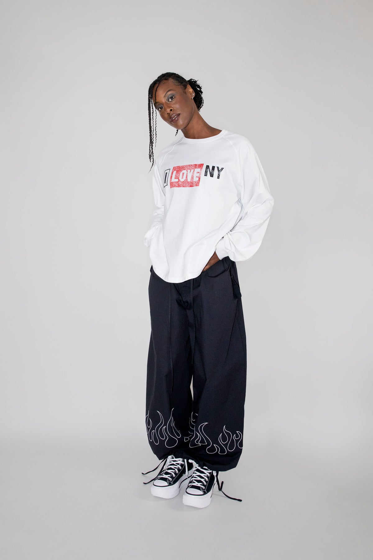 A model stands against a light gray studio background wearing a white long-sleeve crewneck sweatshirt with a red and black 'I LOVE NY' graphic, black wide-leg sweatpants with white flame graphics near the ankles, and black-and-white sneakers.