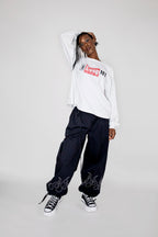 A model posing in a white long-sleeve crewneck sweatshirt with a red and black 'I love NY' logo on the chest, paired with black baggy sweatpants featuring flame graphics near the cuffs, and black and white sneakers, against a light gray studio background.